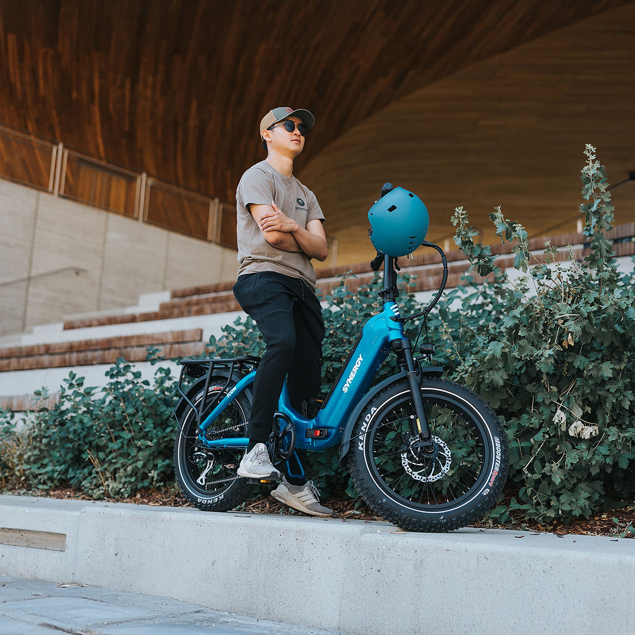 Young man standing with a blue Synergy low-step electric bike in a modern urban park