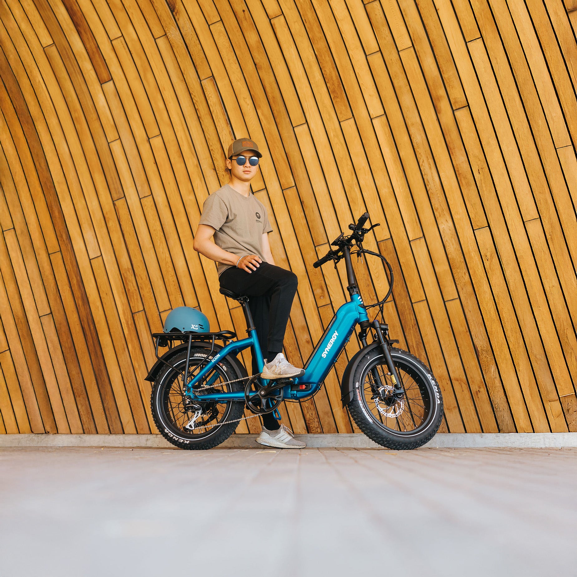 Person sitting on a blue Synergy Icon electric bike in front of a modern wood-paneled wall